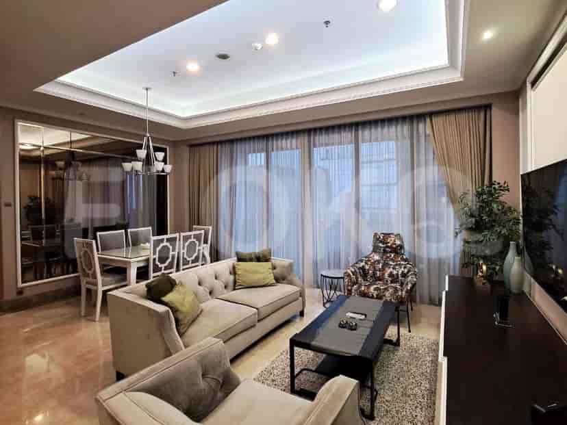 Ruang tamu yang luas dengan sofa abu-abu berbentuk L, TV pintar yang dipasang di dinding, dan jendela besar dengan gorden tipis.