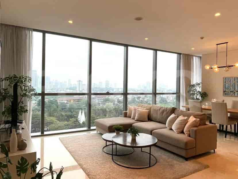 A spacious living room with an L-shaped grey sofa, wall-mounted smart TV, and large window with sheer curtains.