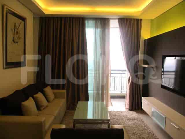 Ruang tamu yang luas dengan sofa abu-abu berbentuk L, TV pintar yang dipasang di dinding, dan jendela besar dengan gorden tipis.