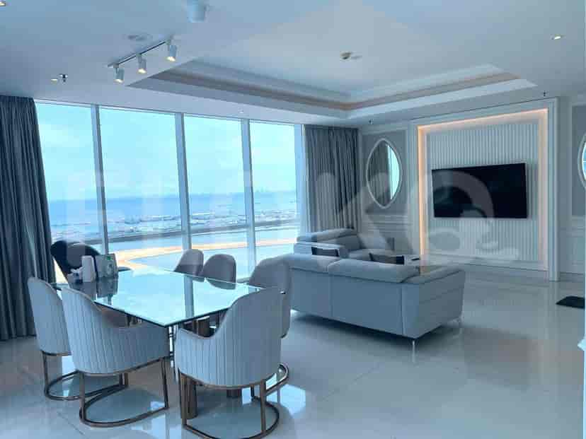 Area ruang tamu dan makan mewah dengan pemandangan laut. Fitur termasuk meja makan kaca, sofa abu-abu, dan TV modern.