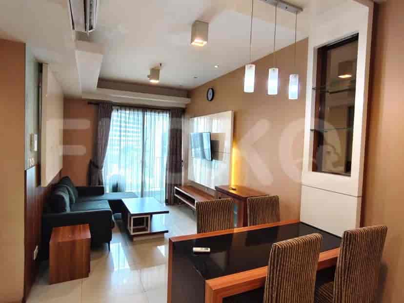 Sebuah ruang tamu modern dengan sofa abu-abu gelap berbentuk L, meja kopi, TV yang terpasang di dinding, dan set makan untuk dua orang.