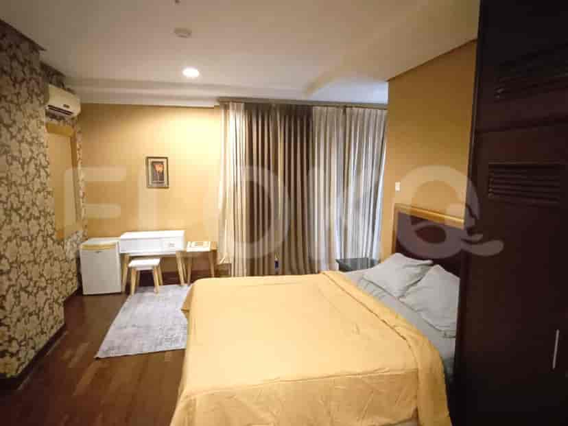 A cozy bedroom featuring a queen-sized bed with yellow bedding, a small white desk with a stool, and a mini-fridge.