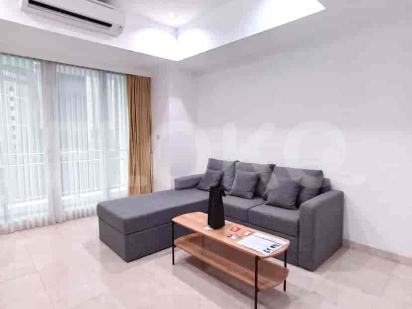 A living room featuring an L-shaped grey sofa, a wooden coffee table with a black vase and some papers, and large windows with beige curtains and sheer drapes.