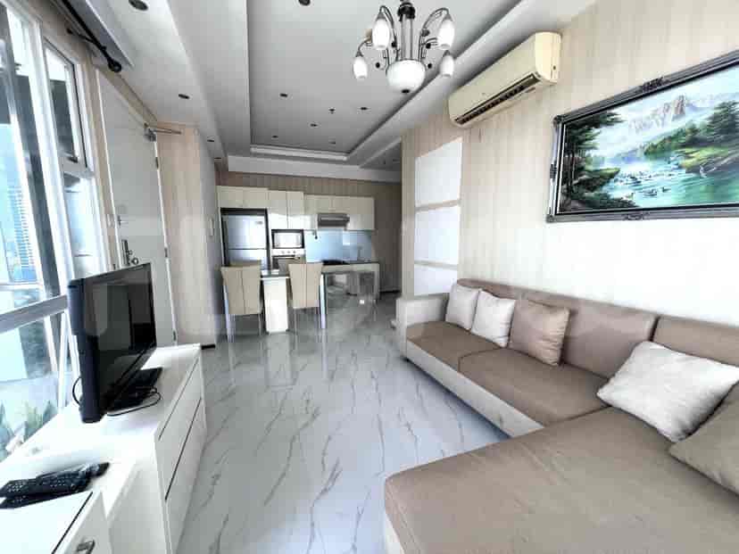 Ruang tamu modern dengan sofa abu-abu, TV layar datar, dan dapur kecil dengan kulkas dan area makan.