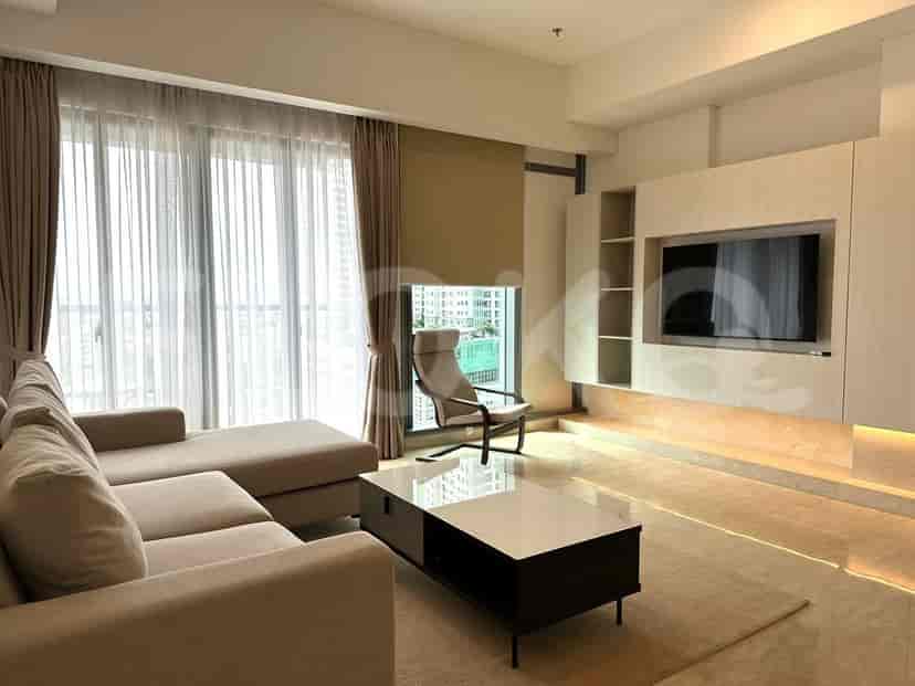 Ruang tamu luas dengan sofa abu-abu berbentuk L, TV pintar yang dipasang di dinding, dan jendela besar dengan gorden tipis.