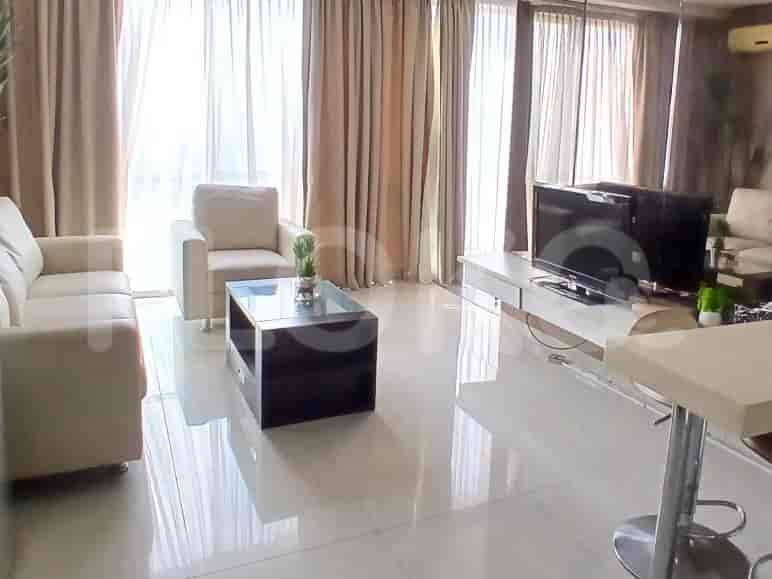 A bright living room with white sofas, a glass coffee table, and large windows with beige curtains.