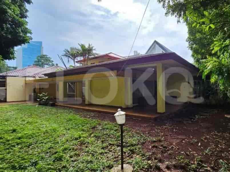 Sebuah rumah kuning dengan teras tertutup, dikelilingi oleh pepohonan hijau subur dan rumput, dengan sekilas bangunan modern di latar belakang.