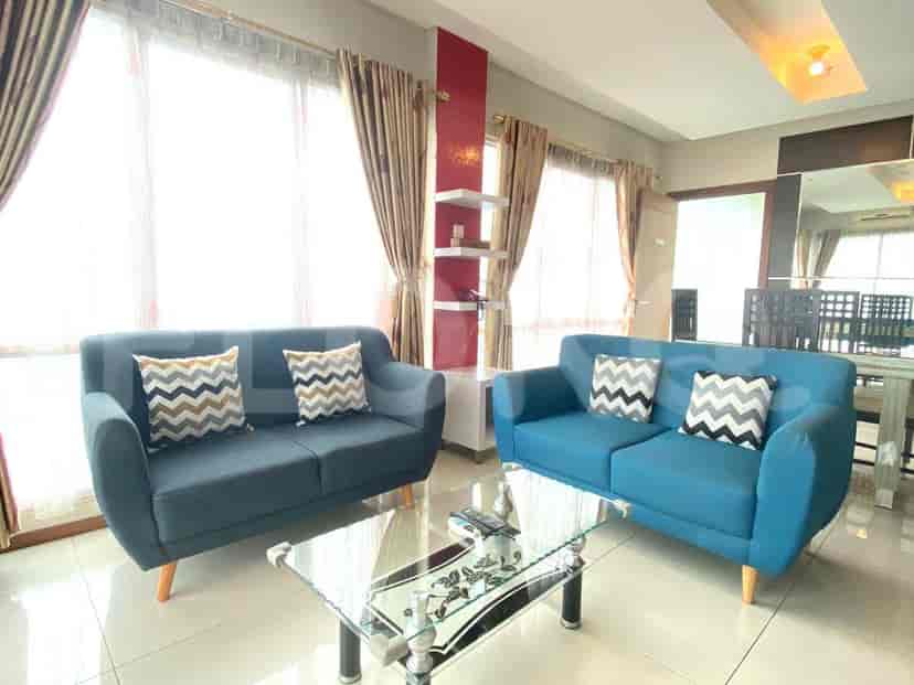 Sebuah ruang tamu modern dengan dua sofa biru, meja kopi kaca, dan jendela besar dengan tirai krem.