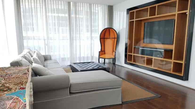 A spacious living room with an L-shaped grey sofa, wall-mounted smart TV, and large window with sheer curtains.