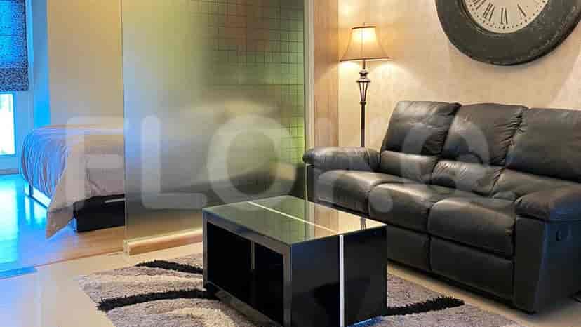 Living room with black leather sofa and glass coffee table, bedroom partially visible behind a glass partition.