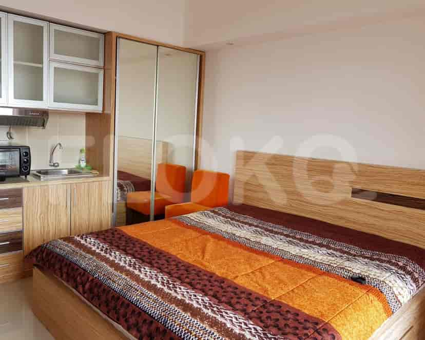 A well-lit bedroom with a bed, small kitchen area, and orange chairs.