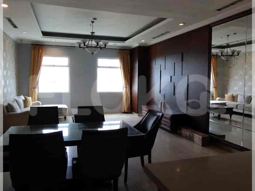 A dining area with a glass-topped table, black chairs, and an attached living area with a cream sofa, windows, and a chandelier.