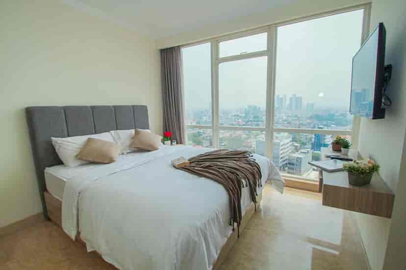 A bright bedroom with a large bed, city view, and a wall-mounted TV.