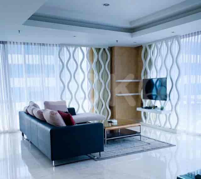 Ruang tamu yang luas dengan sofa berbentuk L, smart TV yang dipasang di dinding, dan jendela besar dengan tirai tipis.