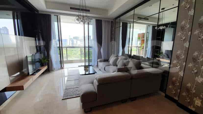Ruang tamu yang luas menampilkan sofa abu-abu berbentuk L, TV yang dipasang di dinding, dan jendela besar dengan gorden tipis yang menawarkan pemandangan kota.