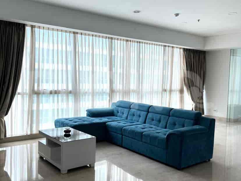 Sebuah ruang tamu modern menampilkan sofa sectional besar berwarna teal menghadap jendela dari lantai ke langit-langit dengan tirai tipis dan gelap, serta meja kopi putih.