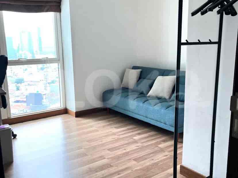 Sudut yang nyaman dengan sofa biru muda, dua bantal putih, dan rak pakaian logam tinggi di dekat jendela dengan pemandangan kota.