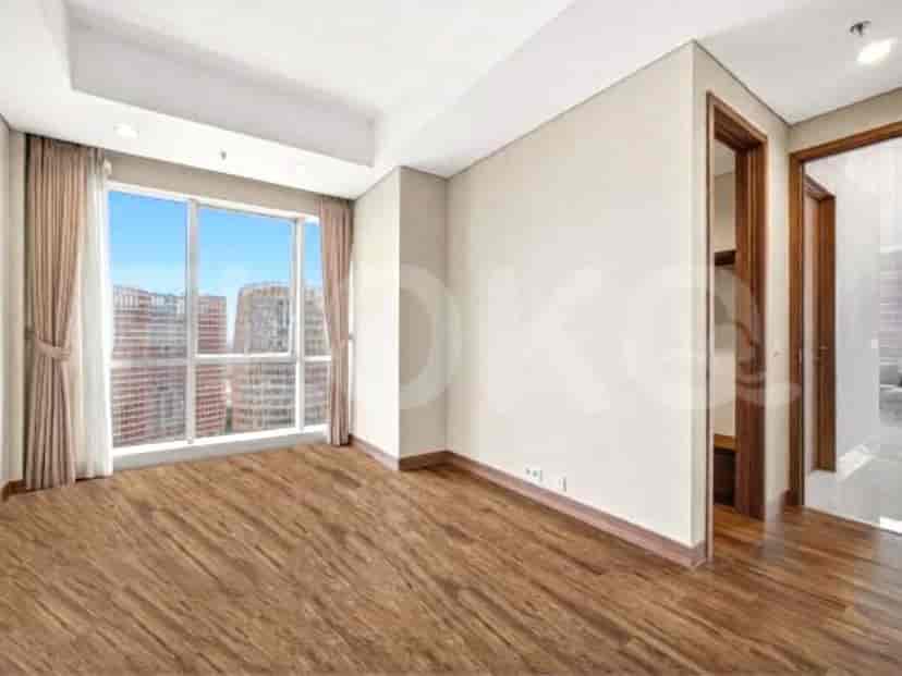 A bedroom with wooden flooring, large windows with beige curtains, and a doorway leading to another room.