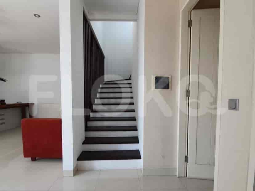 Pemandangan tangga kayu dengan pegangan gelap menuju lantai atas, bersebelahan dengan dinding dengan panel sakelar lampu.