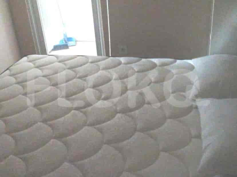 Tempat tidur dengan kasur berpola dan bantal putih, dengan sapu terlihat melalui pintu di latar belakang.